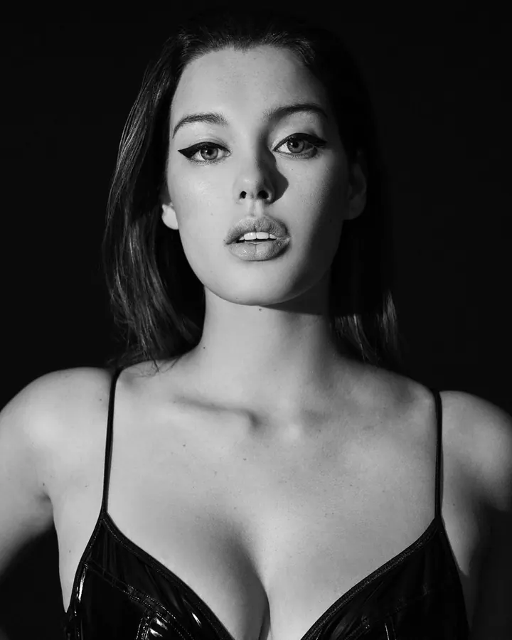 Femme en lingerie noire brillante, photo en noir et blanc, regard intense vers l'objectif.