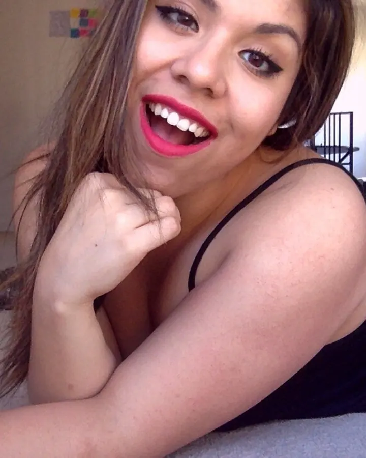 Femme aux cheveux châtains allongée sur le sol, souriant largement avec du rouge à lèvres rose vif, portant un haut noir.