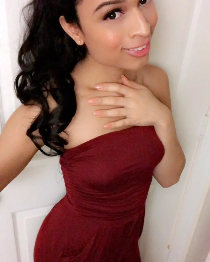 Femme arabe de 18 à 25 ans, portant une robe rouge, souriante, main sur la poitrine, appuyée contre une porte blanche.