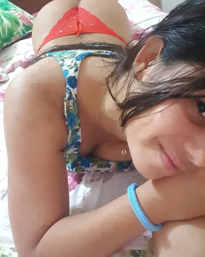 Femme arabe de 18 à 25 ans, portant un haut floral et un string rouge, allongée sur un lit, regardant vers l'appareil, souriante.