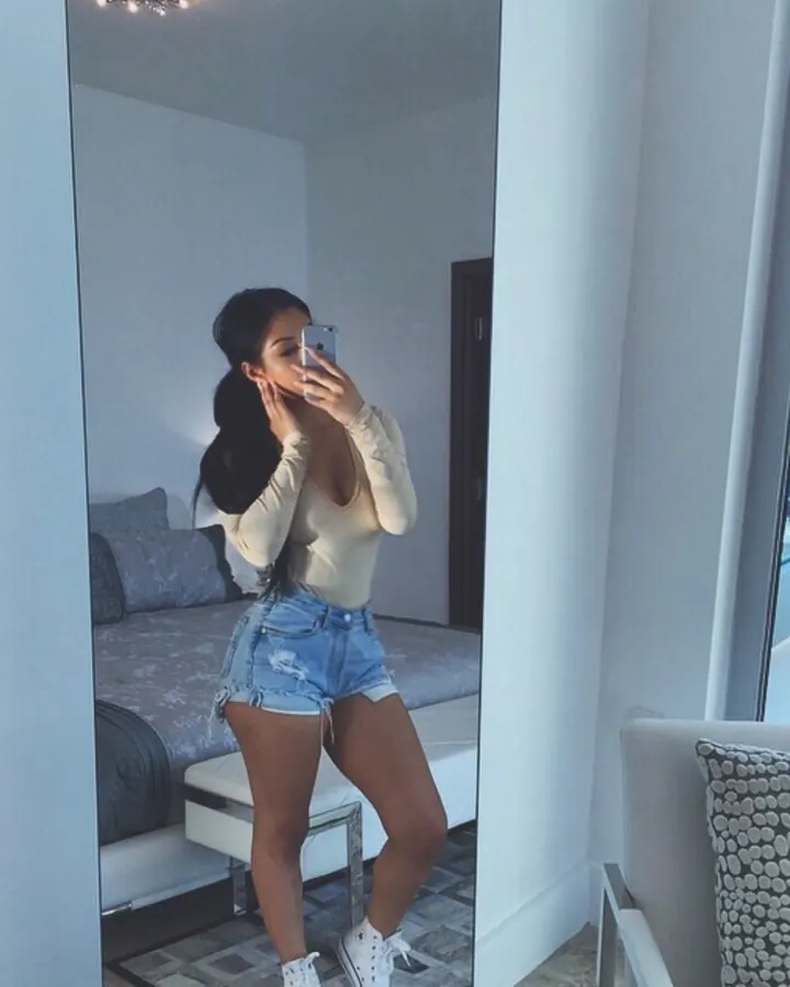 Jeune femme arabe en body beige et shorts en jean, prenant un selfie devant un miroir dans une chambre moderne.
